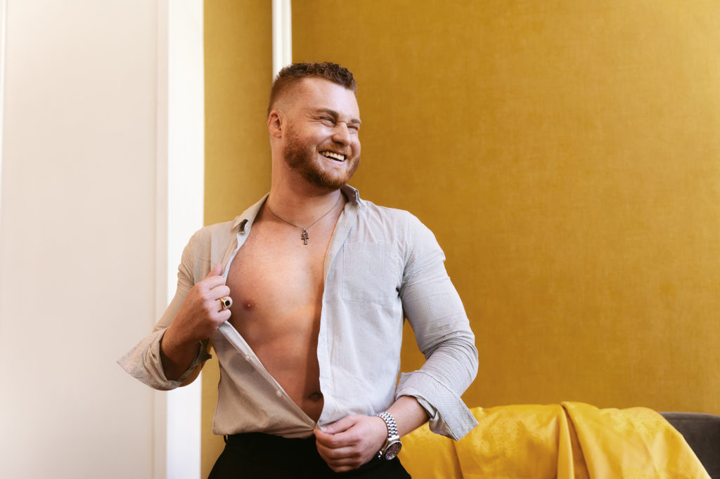Male boudoir session with bearded guy taking shirt off laughing