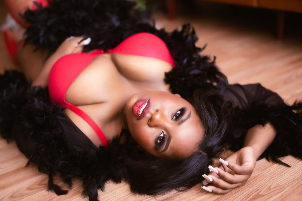 Black woman laying on the floor with red lingerie with black feather robe looking at camera during boudoir session