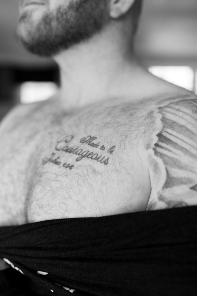 Black and white male boudoir image close up of tattoo on chest hair and beard. Taking off shirt.