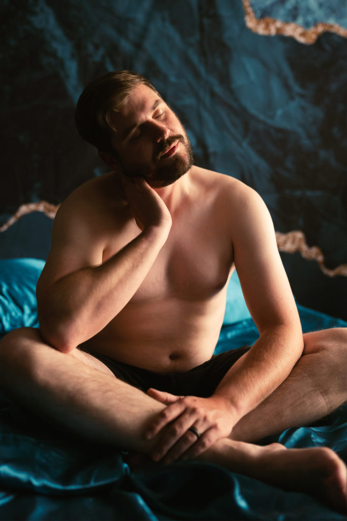 Male boudoir with teal silk sheets in underwear rubbing neck