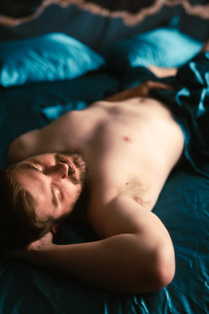 Male boudoir laying on the bed with teal blue sheets shirtless and a beard