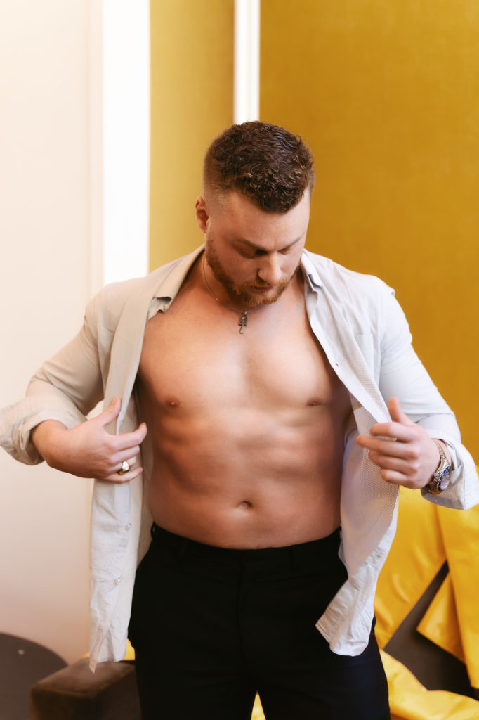 Male boudoir with guy taking off his white button up shirt showing abs with a beard